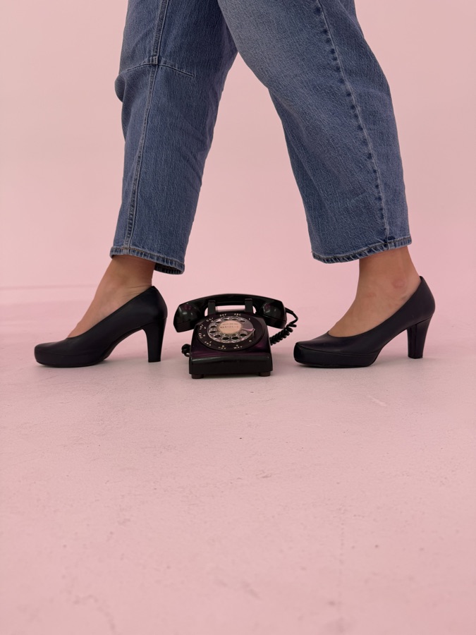 Talons noirs et téléphone vintage posé au sol, sur fond rose